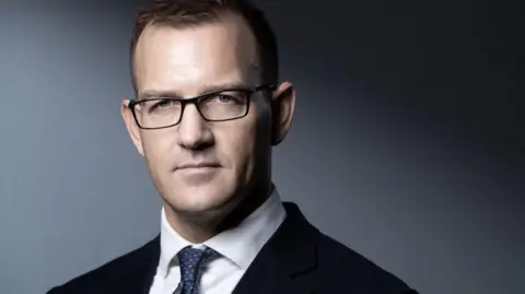 A close up of Daniel Kretinsky in a suit and tie stood in front of a grey wall