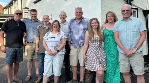 Roxwell Chequers Community Benefit Society A group of nine people in summer shirts and dresses smiling outside The Chequers pub in the evening sunshine.