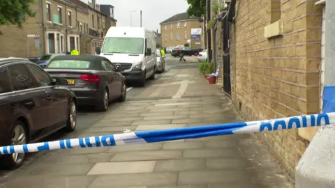 Police tape across a street in Bradford with terrace houses on each side, and two cars and a van parked by the pavement.