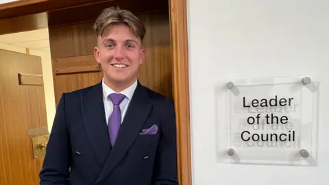 Warwickshire County Council leader George Finch leans against a door frame. The sign to his left reads "Leader of the Council".