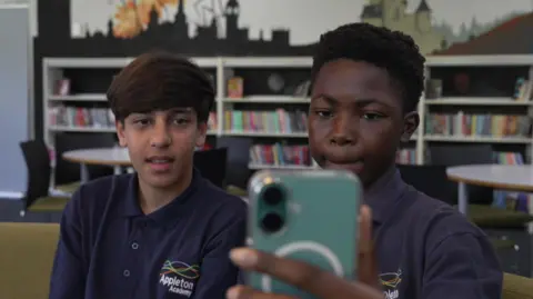 Two boys in school uniform look at a mobile phones. School shelves are in the background. 