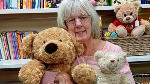 Older woman with round glasses holds three teddy bears, one big, one small, and one medium. Behind her are shelves of books and a teddy bear with a red jumper which has the Wales flag on it.