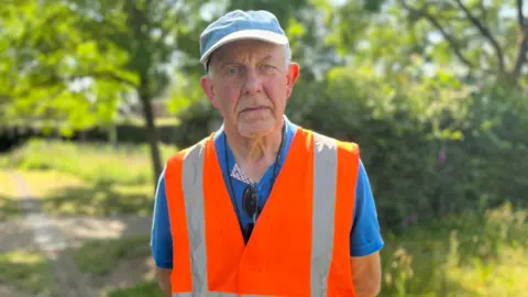 Steve Battle is wearing a blue cap, a bright blue polo shirt and an orange hi-vis jacket. He has a sad expression.