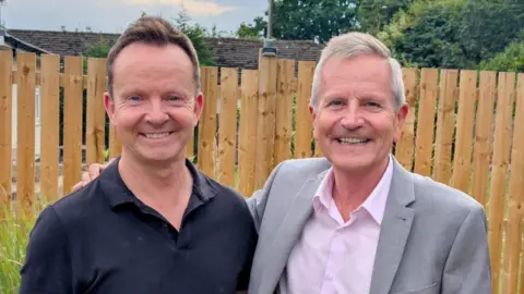 Paul Hudson Man with short brown hair and wearing a dark shirt, standing next to another man with grey hair, who is wearing a pink shirt and grey jacket. The pair are stood in a garden with a wooden-panelled fence in the background.
