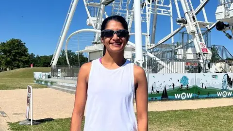 Contributed Rena smiles at the camera in a Milton Keynes park on a summer's day. She has dark hair that is tied up behind her head and wears sunglasses and a white vest top. A ferris wheel can be seen behind her in the park.