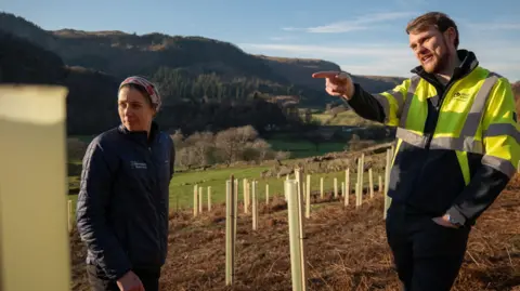 United Utilities West Cumbria Rivers Trust's project manager, Caitlin Pearson, wears dark clothing and stands talking to Andrew Wright, who is wearing a fluorescent United Utilities jacket. They are standing where the trees have been planted.