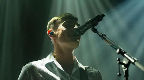 Getty Images A young man sings into a microphone on stage under white strobe lighting, meaning his face is fairly shadowed. The photo is taken from below.