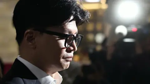 A close-up of Han Dong-Hoon's face, backlit by the flash of a camera