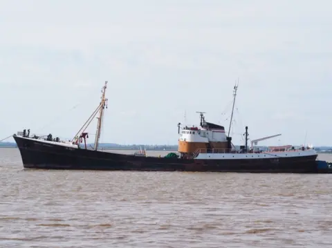 A large ship in water. It has two masts. It is black with a white and brown bridge. There is land in the distance.