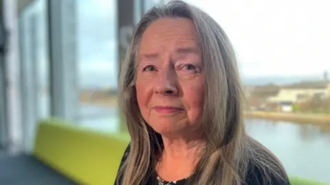 Image of Gina McGavin, a middle-aged woman with long light brown hair. She is sitting on a green sofa with a window running behind her - and a view of the River Clyde.