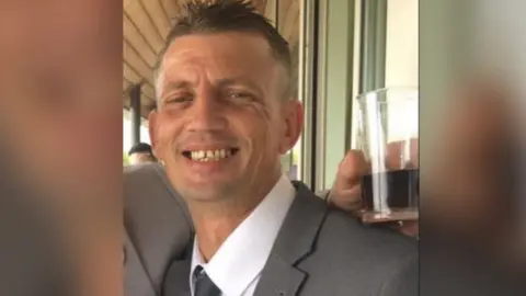 Surrey Police Jason Lucas, wearing a white shirt, grey tie and grey suit, grins into the camera at a social event.