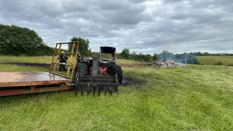 Charred telehandler, with firefighters in the background and small bonfire on field. 