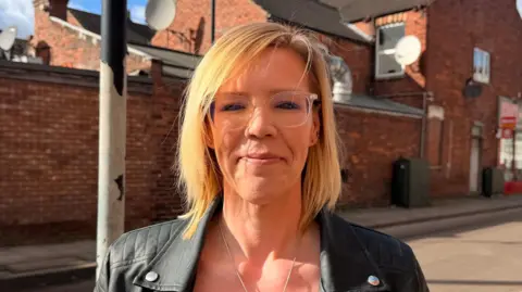 Beckie Stephen stood in the street smiling at the camera. She has shoulder-length, straight, blonde hair and is wearing clear-rimmed glasses, a silver necklace and a black leather jacket. 