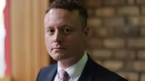 Craig Snee, man dressed in dark blue suit jacket, white shirt and pink striped tie looking at camera