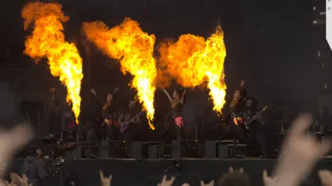 Roger Alarcon/Download Festival Three women on a stage, dancing. There is fire shooting in the air from three flame throwers.