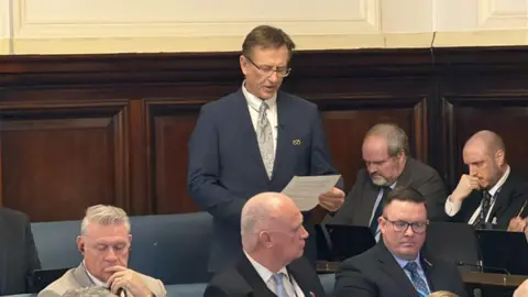 A man standing up in a council chamber holding a sheet of paper. Five other men sit on benches in front and to the side of him