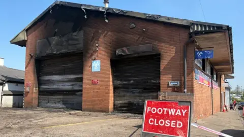 One end of a red brick building with large double garage doors is severely damaged by fire. In front of the building, there is a sign saying 'footway closed'.