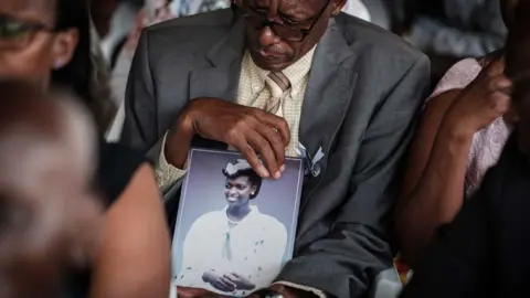 YASUYOSHI CHIBA Vianney Rusanganwa, 61, holds a portrait of his late wife Liberatha Mukangira who died aged 31, during the mass funeral to bury 81 coffins containing newly discoverd remains of 84,437 victims of the 1994 genocide in the mass grave at the Nyanza Genocide Memorial, suburb of the capital Kigali, on May 4, 2019. - The remains of nearly 85,000 people murdered in Rwanda's genocide were laid to rest on May 4 in a sombre ceremony in Kigali, a quarter of a century after the slaughter.