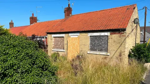 A boarded up bungalow with a very overgrown front garden. The grass is touching the windowsills and a large bush is encroaching on the footpath to the door.