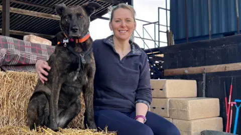 Abbie Condon A woman sat on a straw hay bale cuddling a big dog. She is wearing a quarter zip jumper and smiling.