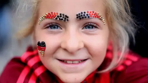 A young girl with blonde hair and blue eyes smiles to the camera in a close up shot. She is wearing sparkly eyebrows made out of beads in the colours of the German flag and a little heart on her right cheek.