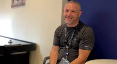 A man in a grey t-shirt smiles widely. He is sitting in a chair that is next to a table. There is an air hockey machine in the background. 