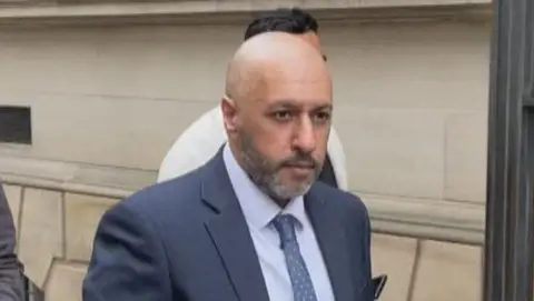 BBC Bald man in a dark blue suit, light blue shirt and blue spotted tied pictured leaving a light sandstone court building. He has a well-trimmed grey beard and is looking in the distance, to the left of the camera. He has a serious expression on his face. 