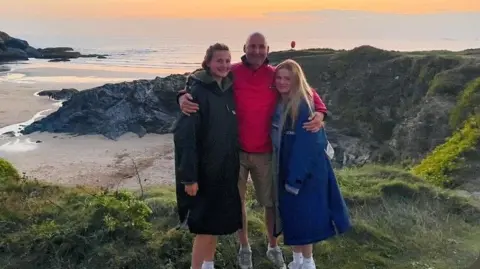 Shaun Lyle A man and two girls stand on the side of a cliff, which overlooks the beach and the sea.
