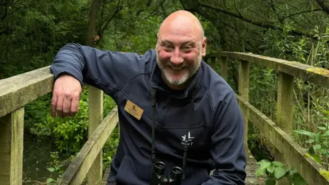 John Gowland is smiling at the camera as he kneels on a wooden bridge at Washington Wetland Centre and resting his arm on the railing. He is wearing navy blue jumper with the white logo of WWT embroidered on it. A pair of binoculars is hanging over his neck. Mr Gowland is bald and has white beard. The bridge is surrounded by lots of trees and grass.