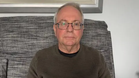 A man, with grey hair, sitting on a grey couch. He is wearing glasses and a dark green jumper.