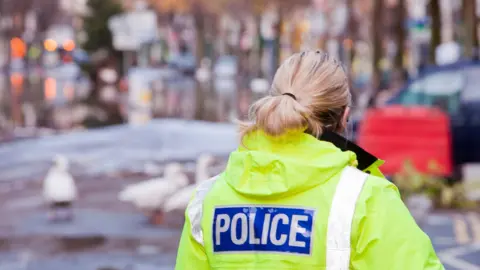 The back of a woman police officer wearing a yellow high vis jacket with the words police written on the back. She is stood in front of a blurred landscape.