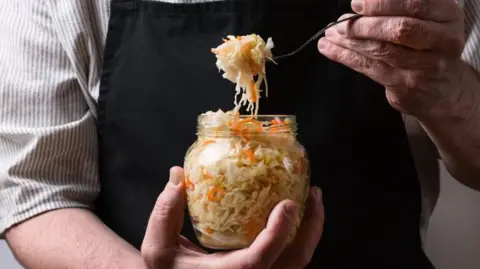 Getty Images A man holds a jar of sauerkraut in one hand and a fork loaded with sauerkraut in another. He is wearing a black apron and a striped shirt. The photo is cropped to only show his chest. 
