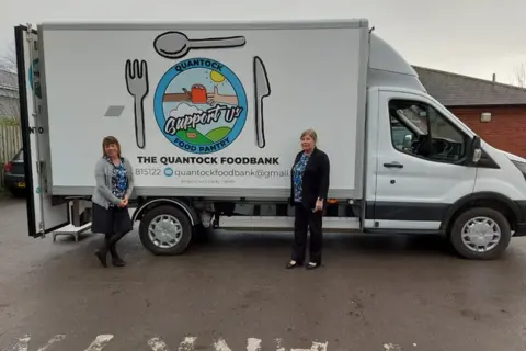 Two women are standing either side of a van with the wording Quantock Foodbank on the side