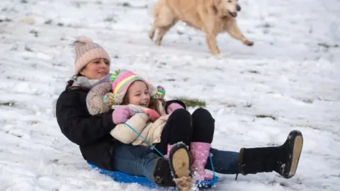 PA Media Lizzy and daughter Connie, 6, are chased down a snow covered hill by their golden retriever Pippa in a park in Newcastle-under-Lyme, Staffordshire.