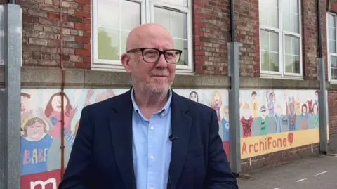 Andy McDonald - a balding man, wearing black-rimmed glasses, a navy blazer and a blue shirt, stands in a school playground. There is a red-brick building with large windows and artwork of children painted on the wall behind him.