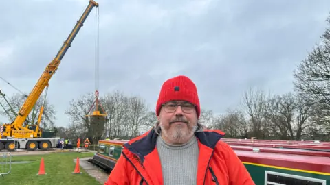 Paul Stowe, a man in a red hat, red jacket and grey jumper i standing next to a line of canal boats. In the background a large yellow crane is visible