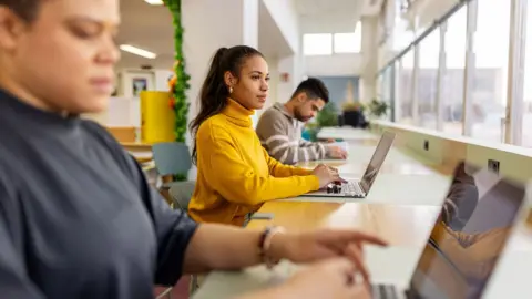Getty Images Stock photo shows students sitting at a table by a window in a student library working away at their computers.