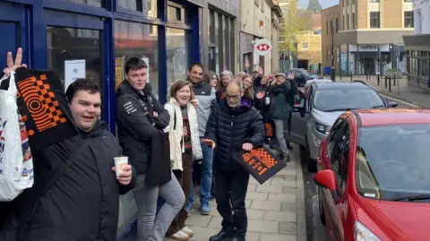 Tom Berry On George Street, people queue on the left-hand side of the pavement in single file. Two people have bags. People are smiling and looking at the camera. 