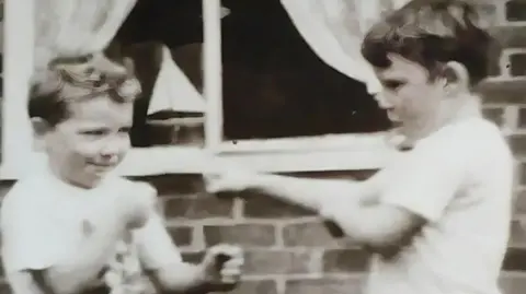 Mike Reid A black and white photo of two young boys standing outside the wall of a house facing each other with their fists raised in a boxing stance. A model sailboat can be seen on the inside of a window ledge behind them. 