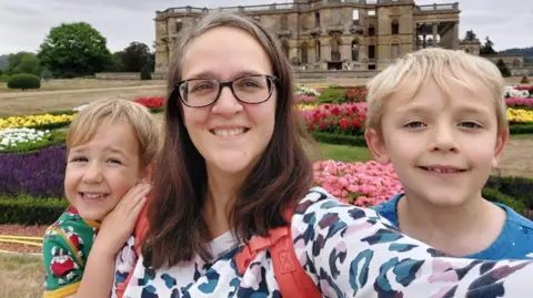 A woman with glasses and long brown hair, greying on top, is standing in front of flowerbeds with what looks like a stately home behind them. On either side of her are two young boys with blonde hair. All three are smiling at the camera and the woman's arm is outstretched to take a selfie.