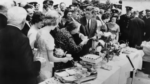 AFP via Getty Images A black and white photo of US President John Fitzgerald Kennedy sharing a lunch with his Irish family during his visit in Dunganstown, County Wexford in 1963. He is wearing a dark suit and tie and is standing at a long table, surrounded by women, while uniformed police stand guard in the background. A woman is handing him a cup of tea. There is a tea tray, a large white cake and flowers on the table.
