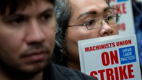 Two people's faces are seen close up alongside a sign that says 'Machinists Union on strike; unfair labor practices!', during strike action in Washington DC on 15 October