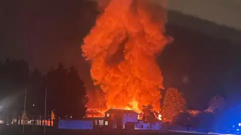 Lucy Groves Picture was taken at nighttime and shows huge clouds of smoke billowing into the sky. The smoke glows red, illuminated by the flames and seems to emerge just behind a small building that sits in front of a large grass area. Several trees surround the fire on either side.