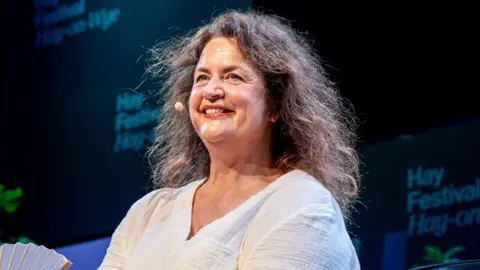 Adam Tatton-Reid/Hay Festival Close-up of Ruth Jones on stage at Hay Festival. She looks out at the audience with a big smile. She has dark brown hair and wears a white linen shirt.