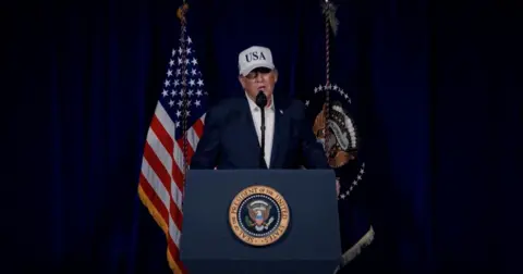 US President Donald Trump speaks at a podium with the US Seal while waring a dress shirt, dark blazer and white hat that is emblazoned with USA.