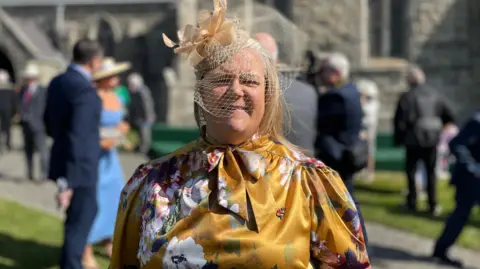 Clare Barber is wearing a yellow floral long sleeved dress with a cream fascinator with netting over her face. She is smiling and there are people milling behind her on a sunny day.