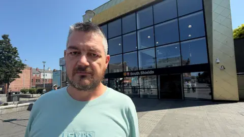 BBC/Jim Scott Mark Berriman is standing outside South Shields Metro station on a sunny, cloudless day. He has short cropped grey hair and a brown and grey beard. He is wearing a greeny-blue t-shirt with a Levi's logo embossed on the front in a slightly different shade. The station is a tall building with the sides and top clad in a gold colour containing symmetrical diamond shapes and surrounding huge darkened windows in a continuous pane of squares marked out with a thin black partition lines. These are arranged in three rows of six above a glass full-width entrance which includes automatic sliding doors.