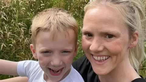 A woman with blonde hair smiles into the camera, she is holding a young boy with blond hair. 