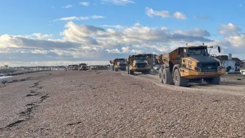 A series of large trucks conducting shingle recycling on the beach between Shoreham and Lancing. There are numerous trucks driving on the beach. 