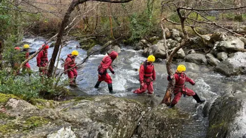 A group of children walking through a rocky river gorge, wearing bright red safety suits, life jackets and yellow protective helmets.